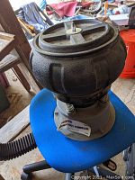 Full view of the vibratory tumbler showing textured black bowl and base on blue chair in a workshop setting.