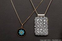 Two silver chains displayed on a dark surface with distinct Murano glass pendants, one rectangular with black and white patterns and one round with blue floral design.