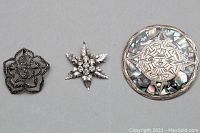 Three silver brooches arranged flat showing the front details: round with abalone and engraved center, star with clear rhinestones, and floral with marcasite stones.