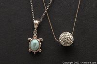 Close-up showing the turtle pendant with oval Larimar stone and the spherical pendant side by side against a black background.