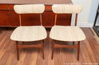 Photo of two teak wood framed Mid-Century Modern dining chairs with light beige striped fabric upholstery on back and seat, showing front view.