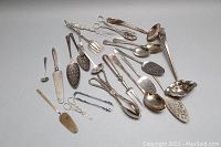 Top-down view of entire lot showing all pieces arranged in a loose grouping, visible are varied silver plated utensils including large ladle, tongs, spoons, and cake servers with visible wear and tarnishing.