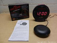 Front view of the Sonic Bomb alarm clock showing digital time display, control buttons, vibrating device, original box, and instruction pamphlet on a table.