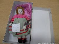 Full view of the Madame Alexander cloth doll inside the original white box with pink tissue paper lining. Doll wears a pink top and green skirt with stripes, red shoes, and has curly brown hair.