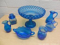 Seven pieces of vintage blue glassware arranged on a table: includes a large pedestal bowl, small pitchers, lidded jar, two-handled bowl, and other small jars/pitchers.