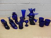 Photo showing entire lot of cobalt blue glass including two bird figurines, vases, boot containers, and multiple drinking glasses.