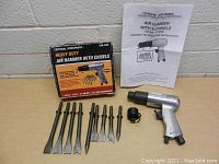 Full set including air hammer, nine chisels, black attachment, instruction manual, and original box against a concrete wall backdrop.