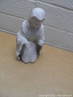 White porcelain figurine of a woman kneeling with head bowed and hands raised, wearing robe, on wooden surface.