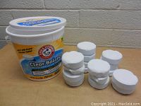 White bucket of Arm & Hammer Clear Balance Clear Pool Tablets with 16 white tablets arranged outside the bucket on a table against a gray wall.