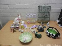 Overview of assorted Easter-themed items arranged on a table under a beige wall. Items include various ceramic bunny and chick figurines, decorative bowls, wire baskets, and bunny ear headbands.