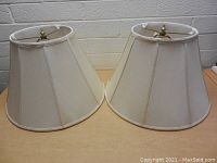 Pair of Ralph Lauren off-white fabric lamp shades with subtle vertical panels and metal harp frames with brass finials on top, showing some even discoloration.