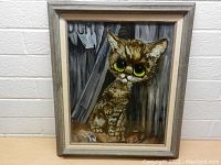 Framed vintage glass cat painting showing cat with large green eyes and a grey curtain background, angled to show frame details.