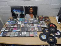 Overview of large lot showing cassette tapes, two 12-inch vinyl LPs, and 7-inch vinyl 45 RPM singles arranged on a wooden table.
