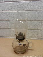 Front view of clear glass vintage hurricane lamp showing glass chimney, metal burner, and round glass base with handle.
