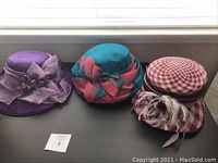 Three woven summer hats displayed side by side on a table near a window: purple with large bow, teal/coral with gathered crown, red/white with feathers and ribbon