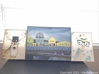 Three art canvases on a shelf by a window, showing all three paintings together including two animal portraits and one village scene.