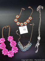 Four costume jewelry necklaces laid out on black surface: bright pink flower bead necklace, blue round beaded necklace, gold-tone necklace with turquoise and coral stones, and plain silver-tone chain necklace