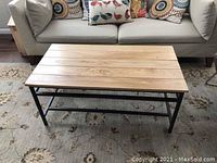 Front angle view of rustic wood plank top coffee table with black metal frame set on patterned rug in front of sofa, showing condition and design.