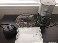 Overview of the lot showing all items including electronic drip coffee maker, cast iron tea pot, glass serving dish and ceramic cheese tray.