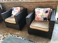 Pair of resin wicker chairs with cushions and floral pillows on patio against brick wall