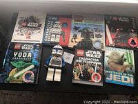 Seven Lego Star Wars themed books displayed on a surface with one large blue and white clone trooper figure in the center. Books include titles about characters, Yoda, and Jedi mysteries.