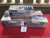 Five RAB DVAKS100CG ceiling mount light fixtures boxed and stacked on a red surface, with visible packaging showing model number and brand.
