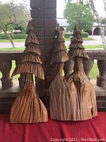 Front view of two carved wooden tree sculptures with layered branches and textured bark, placed on a red cloth with a brick pillar background outside.