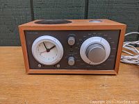 Front view of the Tivoli Audio Model Three clock radio showing the analog clock, volume knob, tuning dial, and speaker grille.