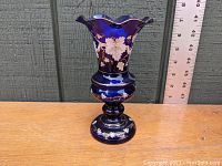 Front view of the cobalt blue blown glass vase with white enamel foliate decoration, showing the flared scalloped rim and pedestal base against a ruler for size reference.