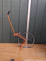 Full view of the orange Rolatape measuring wheel with a wooden backdrop and a board marked in 1-foot increments for scale reference.