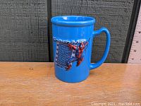 Blue ceramic mug with Spider-Man graphic and informational text, displayed on a wooden surface with a ruler nearby for scale.