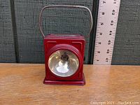 Front view of red rectangular flashlight with large round reflective lens and folding handle on top, showing size reference next to ruler.