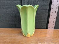 Front view of the six-lobed green pottery vase with a scalloped top and yellowish accents near the base on a wooden surface with a ruler for scale.