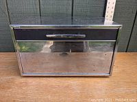 Front view of the chrome bread box, showing the rectangular shape, black trim along the top front edge, and chrome handle. Reflective surfaces with some wear visible.