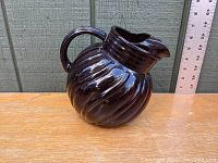 Photo of Royal Ruby Swirl Ball Pitcher showing front-left view with handle and spout, placed on wooden surface with ruler in background for scale.