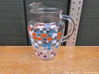 Front and side view of the clear glass pitcher with orange, white, light blue, and purple square pattern resembling TV screen motif. Pitcher has a handle and a pouring spout.