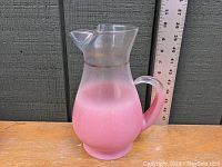 Front side view of mid century pink blendo glass pitcher with clear top and handle shown against a ruler for scale