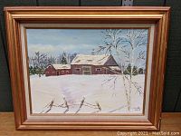 Full view of the framed oil painting showing the snow-covered red barn, trees, fence, and blue sky, with visible signature and frame.
