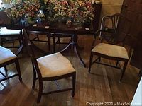 Six wooden dining chairs around a wooden table with floral arrangements on table. Five chairs are armless, one chair has arms and carved shield back. All have upholstered seats.