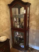 Full view of tall wooden curio cabinet with glass front and sides, filled with glassware and figurines, corner placement near floral wallpapered wall.