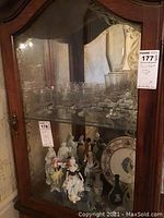 Full view of cabinet glass shelves showing various glasses, porcelain figurines, plate, and vase from a distance.
