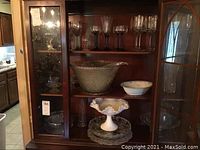 Wide shot showing upper shelves of china cabinet containing various glassware: punch bowl, punch cups, cake plate with cover, milk glass compote, and candle holders.