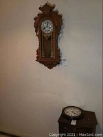 Photo showing two clocks setup: wooden pendulum wall clock mounted on wall, Ingraham round quartz clock lying on a small table.