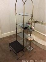 Wide angle photo showing tall gold bamboo style metal shelf with glass shelves, smaller triangular metal and glass shelf unit, and small black metal table in bathroom corner