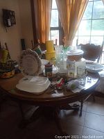 Wide view of all items including ceramic bowl, glassware, Tupperware, kitchen tools on wooden table near window with yellow curtains.