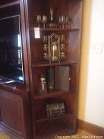 Overall view of wooden shelving unit containing all items in the lot including decanter, glasses, metal stand, bottle, case, and wood chest.