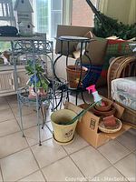 Two metal plant stands and multiple clay pots arranged in a sunroom with some boxes and furniture.