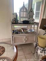 Full view of white rolling cart with birdcage, birdhouses, pots, and frames on and around it near window