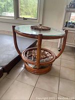 Side angle view showing round glass top and lower woven rattan shelf on tiled floor.