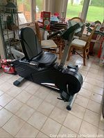 Full side view of the black FitnessQuest Edge 491 recumbent exercise bike showing padded seat and pedals on tiled floor in a sunroom.